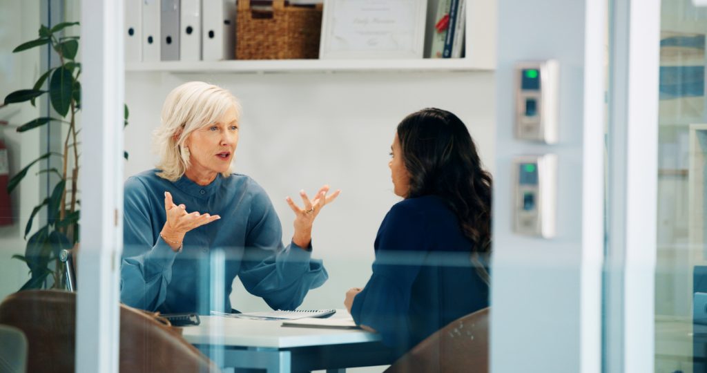 Duas mulheres em reunião em uma sala de escritório envidraçada, sentadas frente a frente, conversando enquanto uma delas gesticula.