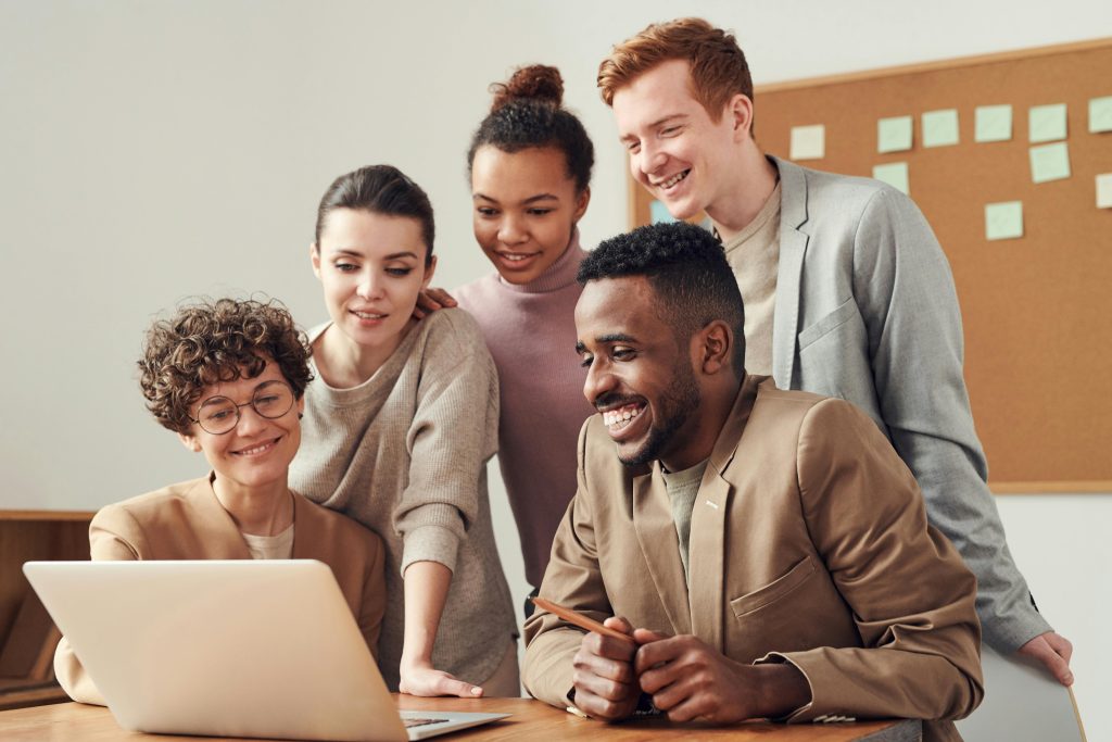 Grupo de cinco pessoas reunidas em volta de um notebook, sorrindo e colaborando em um ambiente de trabalho.