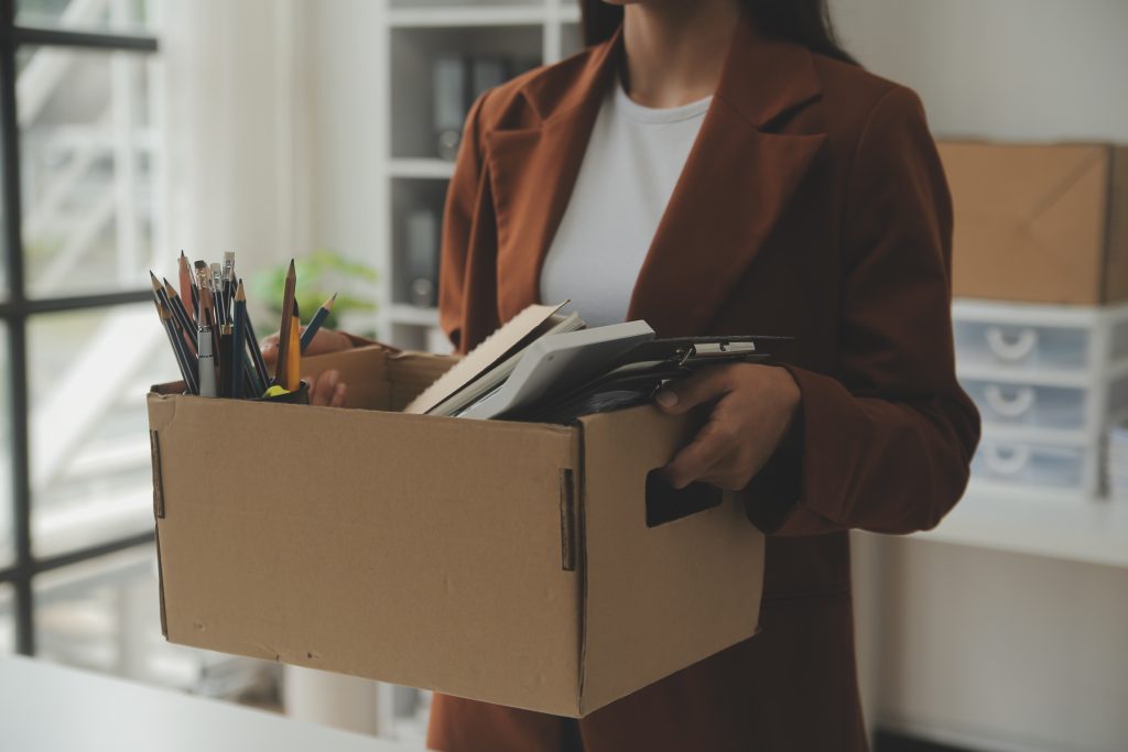 Pessoa segurando uma caixa de papelão com materiais de escritório, como cadernos e lápis, em um ambiente de trabalho.