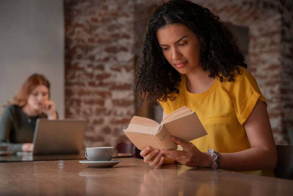 Uma mulher de cabelos cacheados e blusa amarela lê um livro em um café, com uma xícara à sua frente e outra pessoa ao fundo usando um notebook.