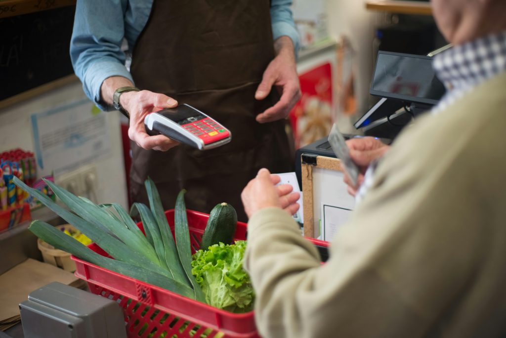 Cliente realiza pagamento com cartão em um mercado enquanto o atendente segura a maquininha; a cena representa a importância da segurança digital nas transações comerciais.