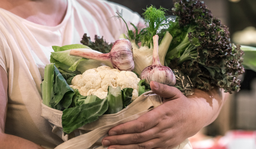 Pessoa segura sacola reutilizável com verduras, couve-flor, alho e outros alimentos frescos.