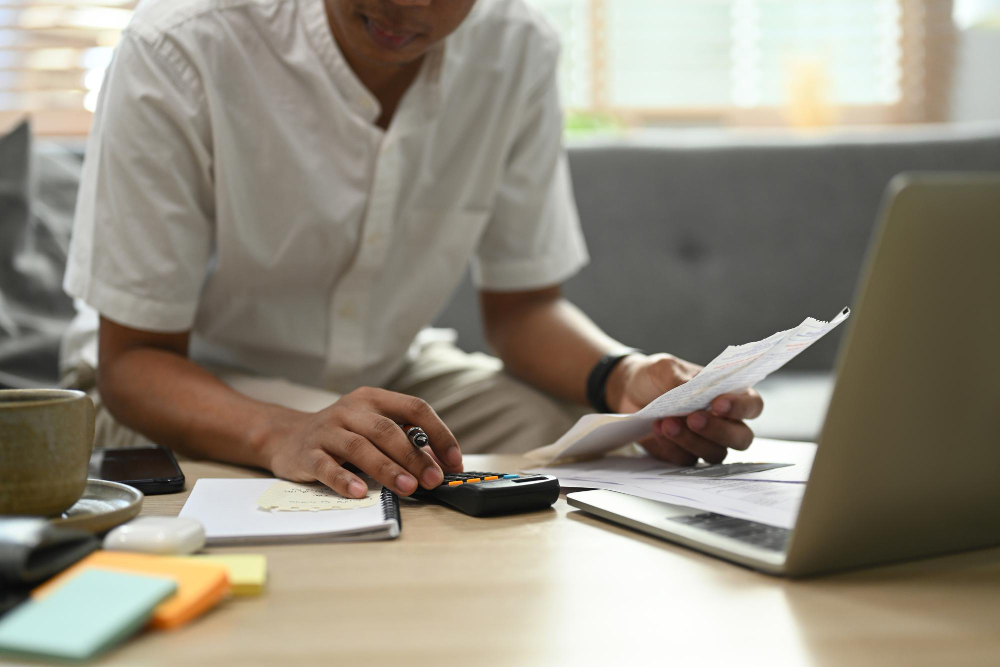 Pessoa sentada à mesa, analisando uma conta e fazendo cálculos com uma calculadora ao lado de um notebook.